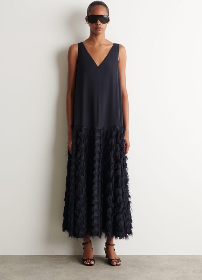 Woman wearing black sunglasses and a sleeveless black dress with a V-neckline and textured, feathered skirt, standing against a plain white wall. Showcasing high street fashion, she completes the chic look with black open-toe heels.