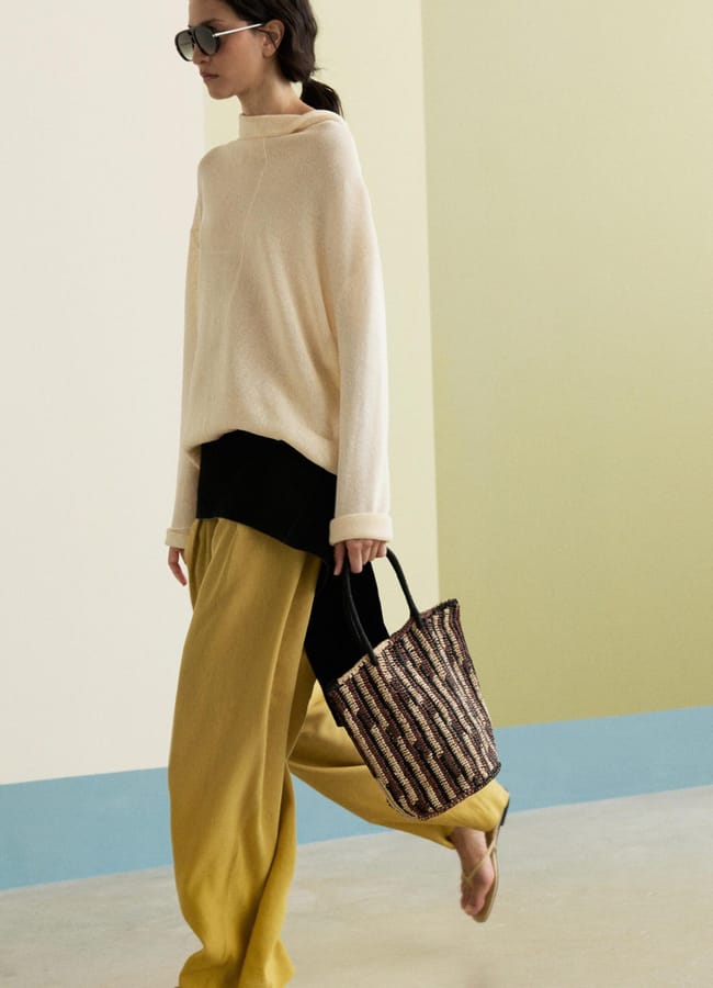 A person embodying high street fashion wears sunglasses, a loose cream jumper, yellow wide-leg trousers, and flip-flops while walking indoors with a woven tote bag. The background is minimal with pastel colours.