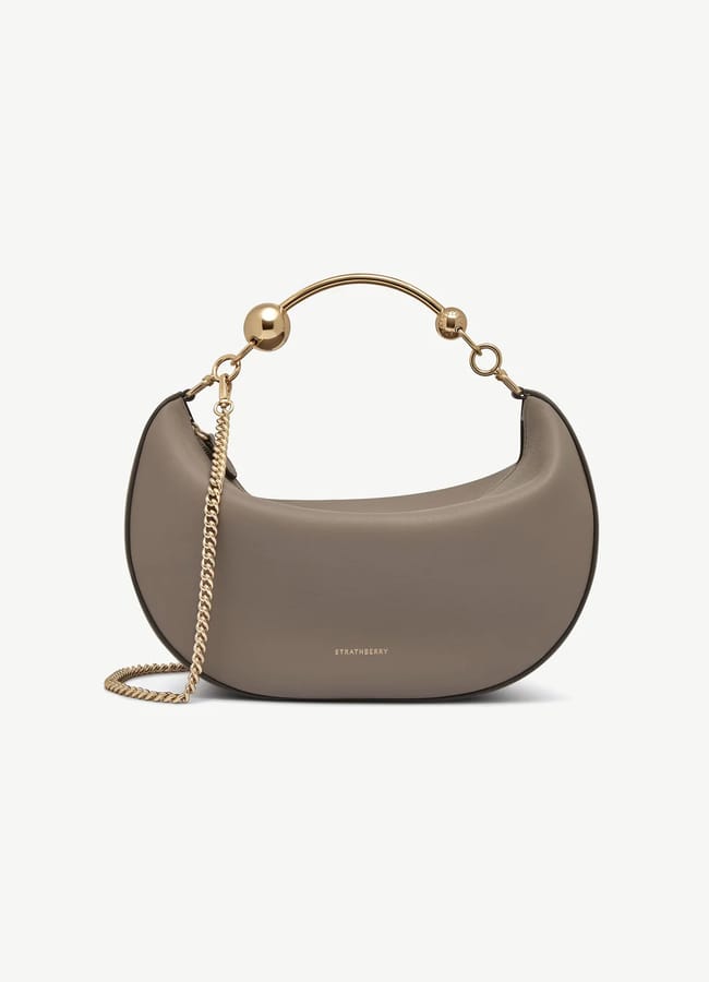 Taupe crescent-shaped designer bag with gold handle and chain strap, featuring a minimalist design and small gold logo on the front—this high-end accessory exudes the understated elegance of a fashion editor’s favourite, set against a plain white background.
