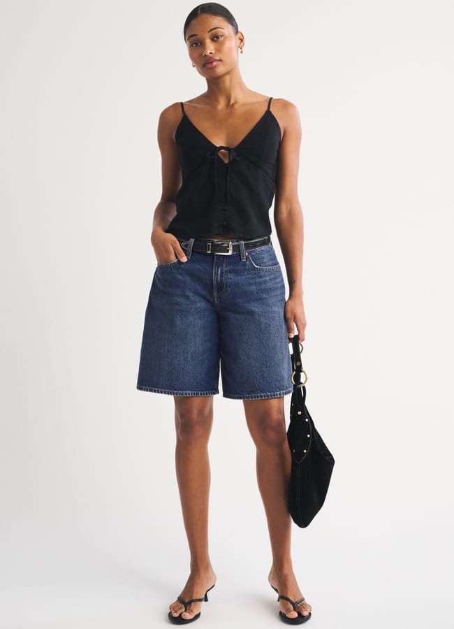 A woman stands against a plain background wearing a black sleeveless top, best denim shorts at knee length, black sandals, and holding a black handbag. She has short hair and a neutral expression.