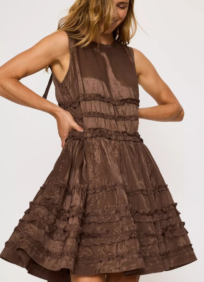A woman in a sleeveless, knee-length, brown ruffled dress stands against a plain background, looking down and smiling with her hands at her waist—radiating effortless high street fashion charm.