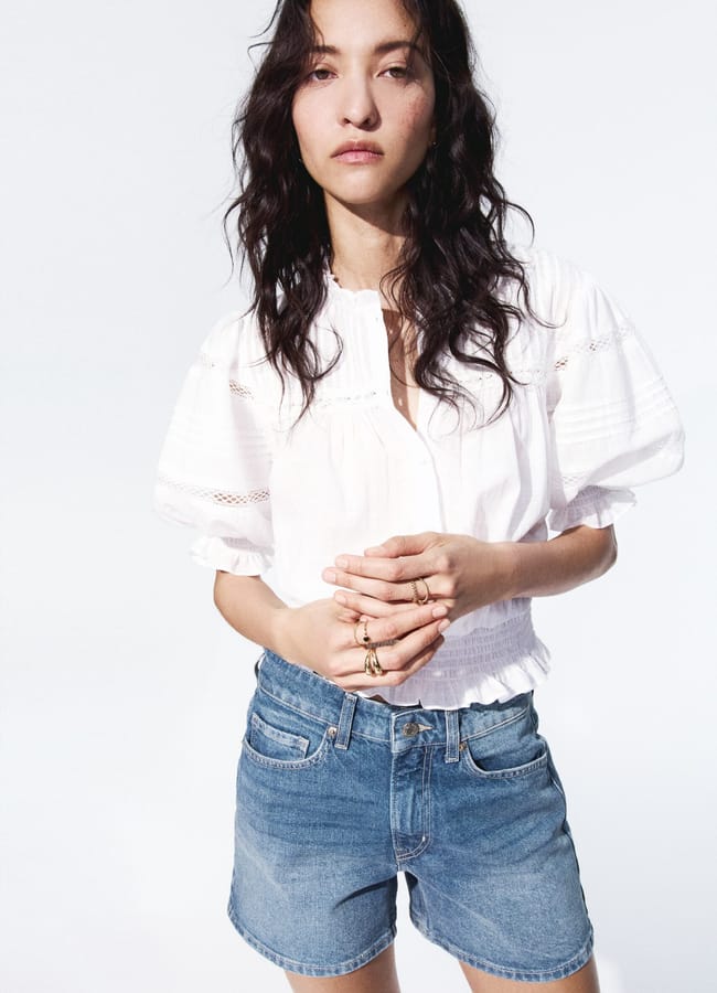 A woman with wavy dark hair wears a white short-sleeved blouse and the best denim shorts, standing against a plain white background with her hands clasped together.