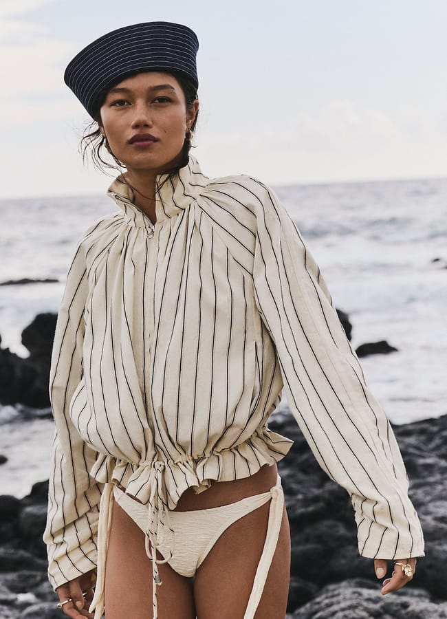 A woman stands on a stony beach, embodying summer micro trends in a striped, long-sleeved, oversized shirt with drawstrings and cream bikini bottoms. She also wears a navy striped hat, with the sea visible in the background.