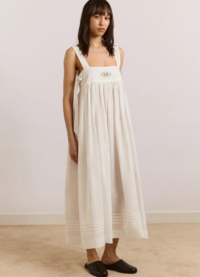 A woman stands indoors wearing a long, sleeveless white dress with wide straps, subtle embroidered detail—a perfect nod to summer micro trends—paired with black slip-on shoes. She has straight, dark hair with a fringe and a neutral expression.