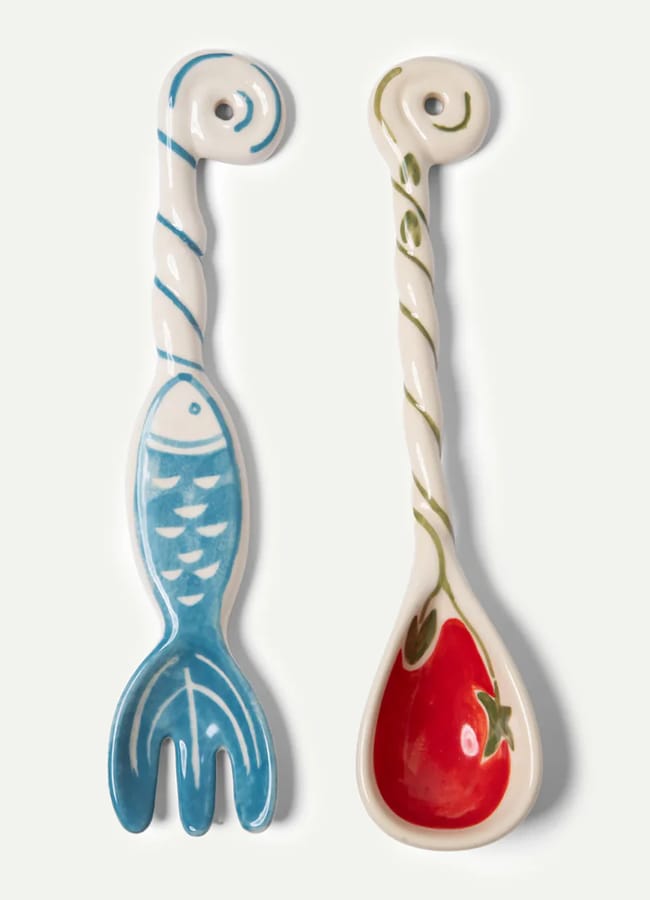 A pair of ceramic utensils perfect for salad season: a blue fish-shaped fork and a white spoon with a red tomato design, both featuring spiral handles and decorative painted details.