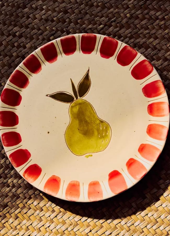 A ceramic plate with a painted green pear and brown leaves in the centre, surrounded by a red border, sits on a woven brown mat in warm lighting—an elegant piece that brings charm to any collection of gourmet cookwear.
