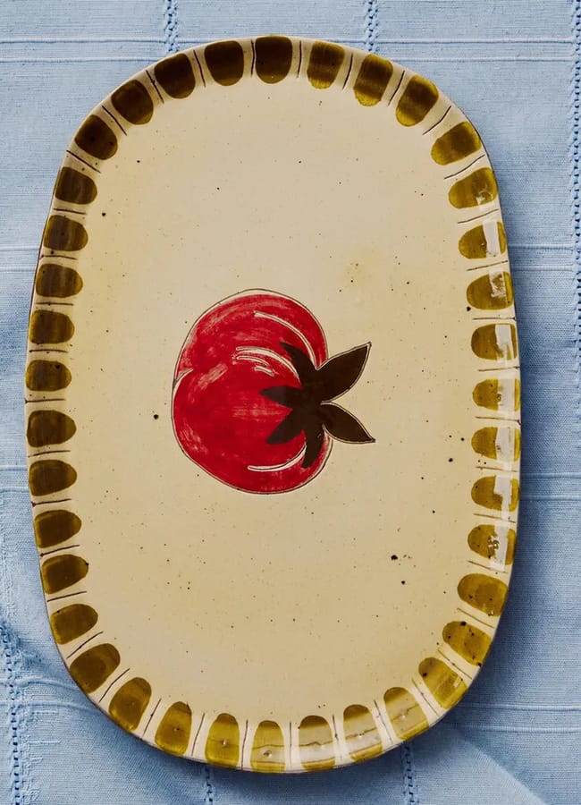 A rectangular ceramic plate with a yellowish border and a red tomato illustration in the centre, perfect for any foodie, is placed on a light blue fabric with stitched lines—an elegant addition to your homewares collection.