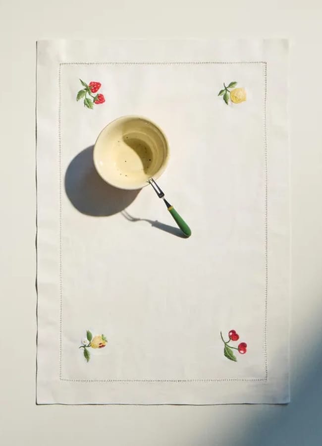 A cup of coffee with a green-handled spoon rests on a white cloth featuring embroidered strawberries, cherries, and a yellow rose at the corners, all set against a plain background—a charming scene sure to delight any foodie or lover of unique homewares.