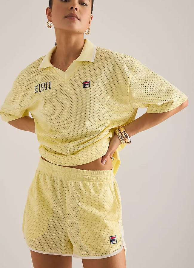 A person wearing a light yellow mesh polo shirt and matching shorts with Fila logos and 1911 text on the shirt, posing with one hand on their hip in front of a neutral background—a fresh take on summer fashion that stands out as one of the top denim alternatives for warm-weather style.