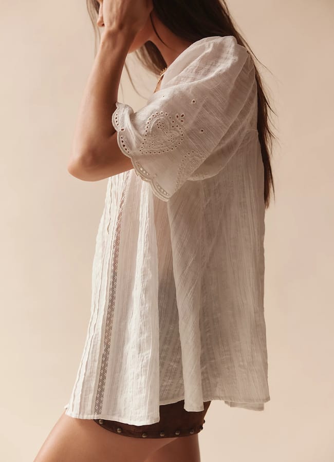 Woman wearing a white, loose, short-sleeve blouse with broderie anglaise details and brown shorts, standing against a neutral background, with her hand touching her hair—an effortlessly chic look that captures the essence of high street fashion.