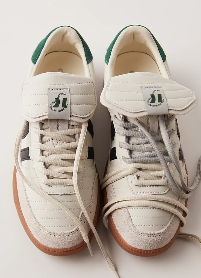 Two white trainers with gum soles, green heel accents, and unique tongue tags showcase an Unlikely Trainer Trend. The left shoe features beige laces, while the right shoe sports grey laces. Both shoes are positioned side by side on a white background.