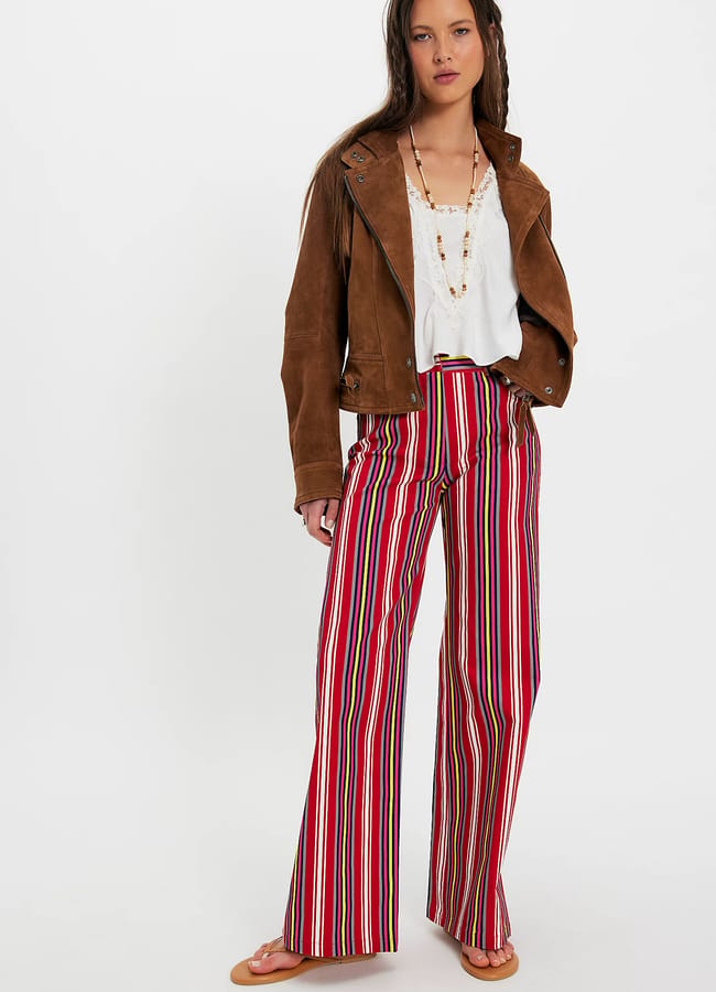 A woman stands against a white background wearing a brown suede jacket, a white blouse, multicoloured striped wide-leg trousers—a nod to summer micro trends—beige shoes, and a beaded necklace. She has long brown hair and a relaxed expression.