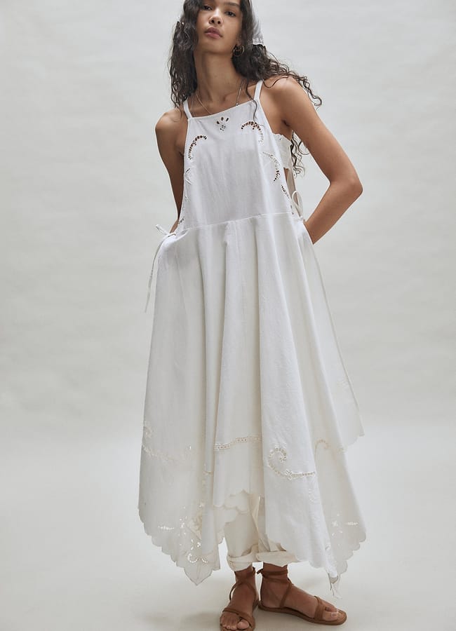 A woman with long, wavy hair wears a flowing, white, sleeveless dress with delicate embroidery—an effortless nod to summer micro trends—layered over loose white trousers and tan sandals, standing against a plain, light background.
