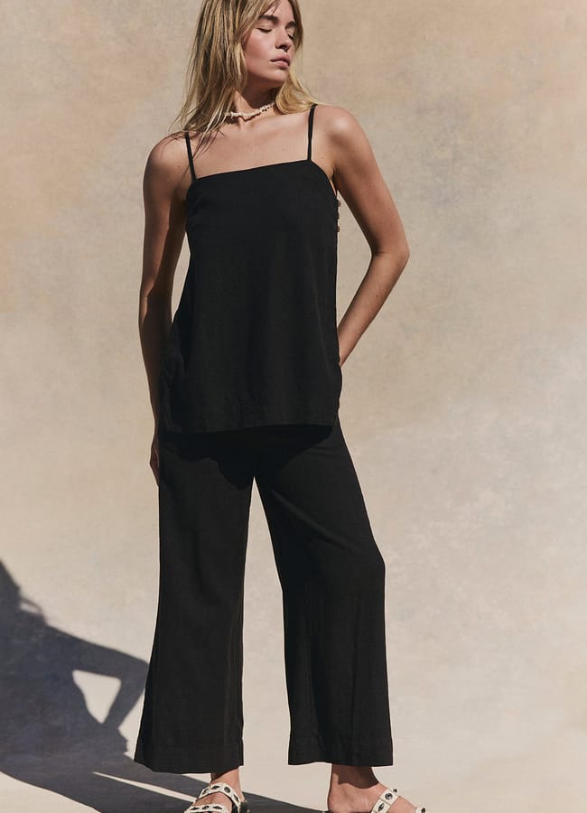 A person stands in a loose black sleeveless top and matching wide-leg trousers, wearing sandals and a necklace—an effortlessly chic choice perfect for summer holiday outfits—against a neutral background.