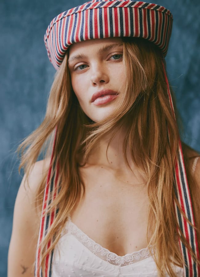 A woman with long light brown hair wears a white lace camisole and a red, white, and blue striped hat with long ribbons—an eye-catching nod to summer micro trends—standing in front of a blue textured background.