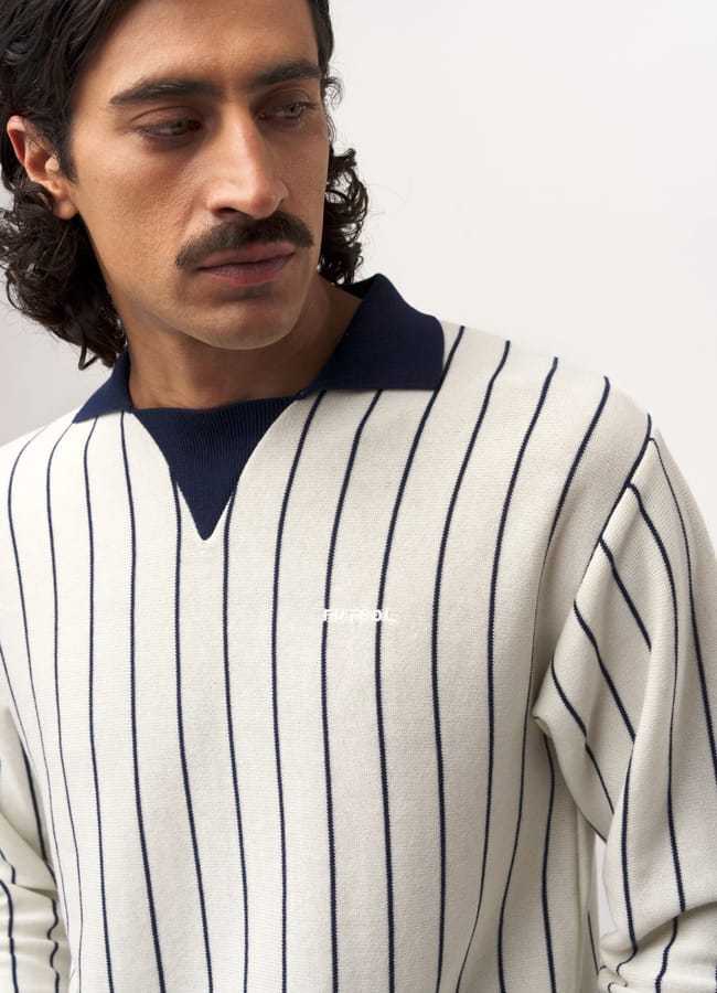 A man with wavy dark hair and a moustache looks down, wearing a cream-coloured, long-sleeve shirt with vertical navy blue stripes and a dark collar—a subtle nod to football core style.