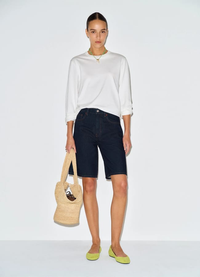 A woman stands against a white background wearing a white long-sleeved top, best denim shorts in a dark knee-length style, light green flats, and a green necklace. She carries a woven straw bag and has her hair tied back.
