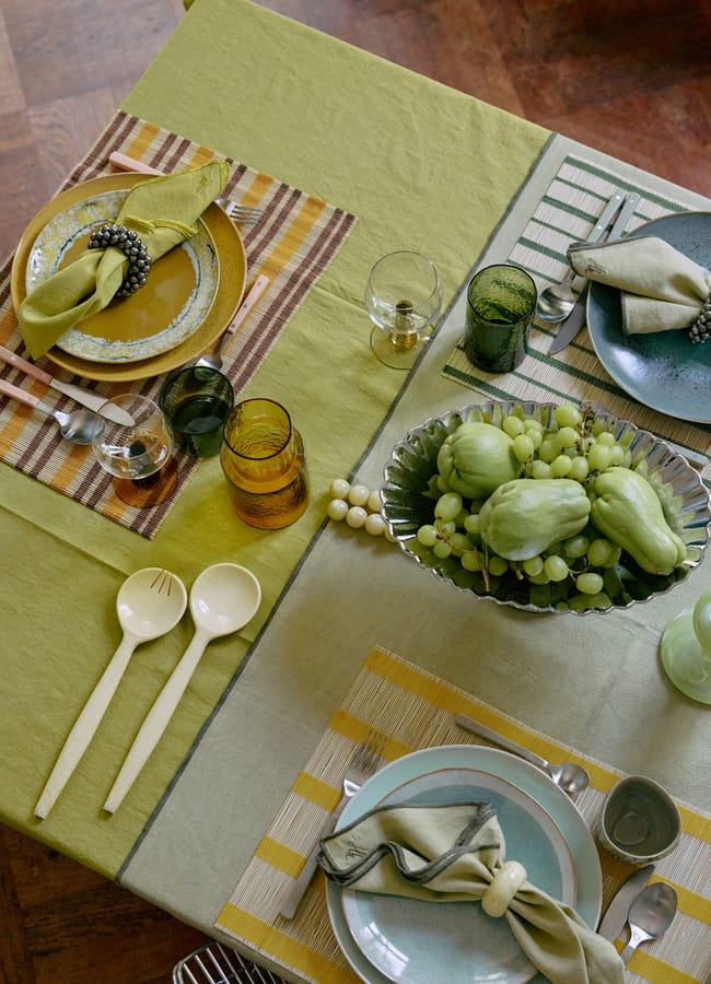 A table set for three with green and yellow tones, perfect for salad season, features plates, cloth serviettes with rings, glasses, and a centrepiece bowl of grapes and green vegetables on striped table mats.