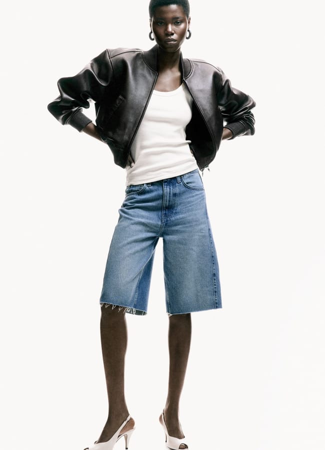 A person stands confidently wearing a black leather jacket, white vest, best denim shorts at knee length, and white high-heeled shoes against a plain white background.