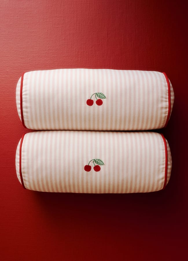 Two cylindrical cushions with pink and white stripes, each featuring an embroidered pair of cherries and green leaves, placed side by side on a red textured surface—a delightful addition to any foodie-inspired homewares collection.