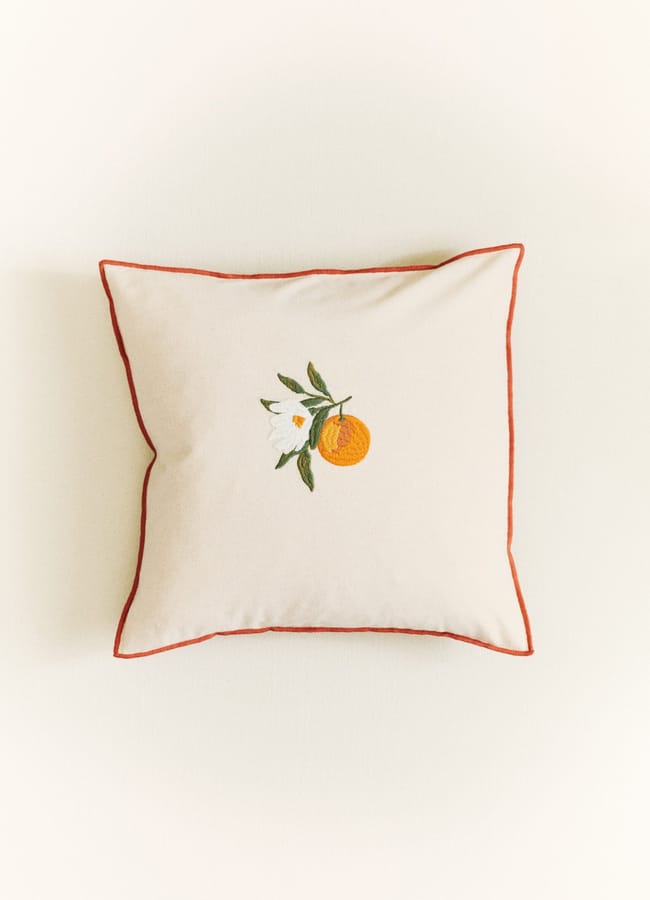 A square beige cushion with orange piping and an embroidered design of a white flower next to an orange fruit at the centre, this charming accent is perfect for adding a touch of foodie flair to your homewares collection.