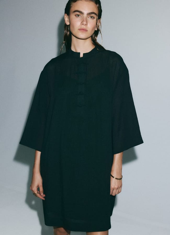 A person with wet, slicked-back hair wears a loose, knee-length black dress with wide sleeves and a mandarin collar—an effortlessly chic outfit that highlights subtle fashion detail—standing against a plain light grey background.