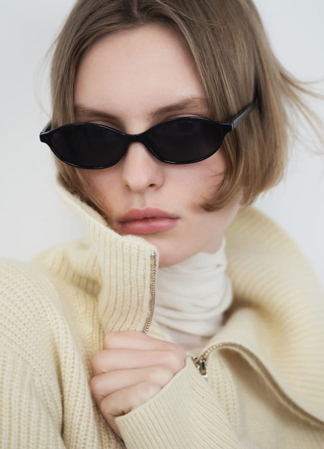 A person with short, light brown hair wears black cat-eye sunglasses, a cream zip-up jumper, and a light polo neck. Channelling Kendall Jenner style, they look away from the camera, holding the jumper collar up with one hand.