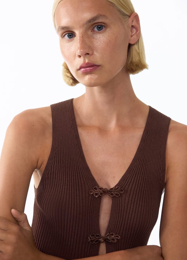 A person with blond hair and light skin wears a sleeveless, ribbed brown top with two decorative fastenings on the front—a subtle fashion detail that adds interest to the look. The background is plain white.