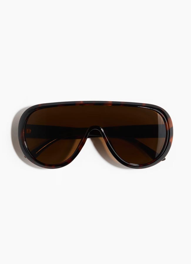 Brown-tinted, oversized shield sunglasses with a tortoiseshell frame, shown against a white background—an effortlessly chic accessory and the perfect answer to what to wear in Ibiza.