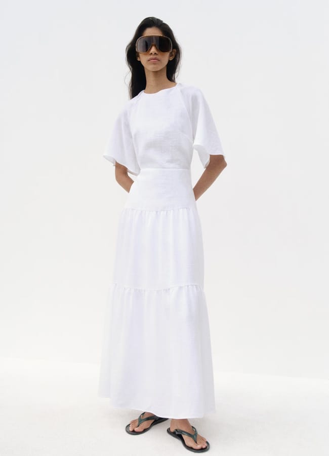A woman wearing a white short-sleeve blouse and a long white skirt stands against a plain background, perfectly capturing what to wear in Ibiza. She has long dark hair, wears large black sunglasses, and black flip-flops.