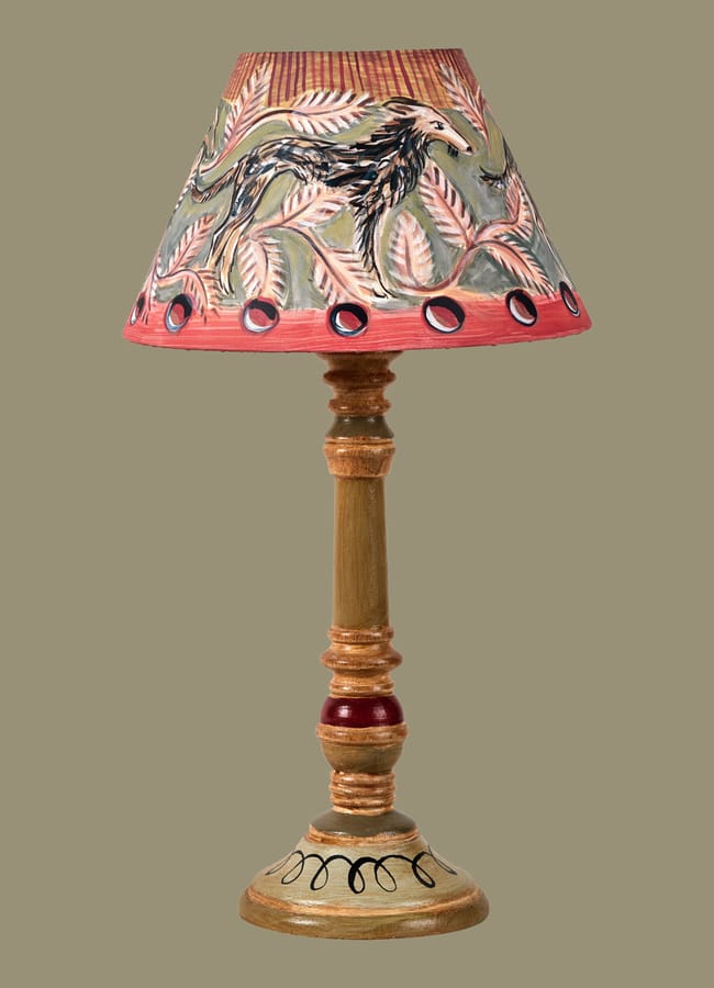 A wooden table lamp with a hand-painted lampshade featuring leaves and a black bird, accented with red and green patterns on a plain tan background, makes an eye-catching statement perfect for maximalist hotels.