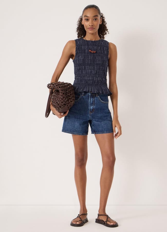 A woman stands against a plain background wearing a sleeveless navy textured top, best denim shorts, and black sandals. She carries a large woven brown bag and has curly hair tied back.