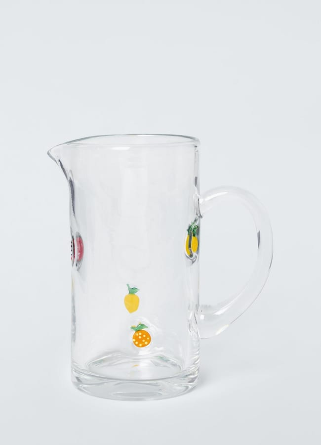 A clear glass jug with a handle, decorated with small colourful fruit illustrations—including lemon and orange slices—against a plain light background, makes a charming addition to any collection of kitchen accessories or foodie homewares.