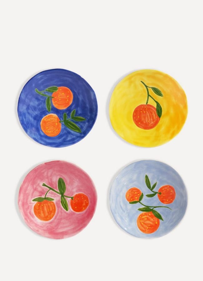 Four ceramic plates, each with a hand-painted orange fruit and green leaves, sit on different coloured backgrounds—blue, yellow, pink, and light blue—arranged in a grid on a white surface. Perfect as unique kitchen accessories or thoughtful cooking gifts, these vibrant dishes add charm to any foodie homewares collection.