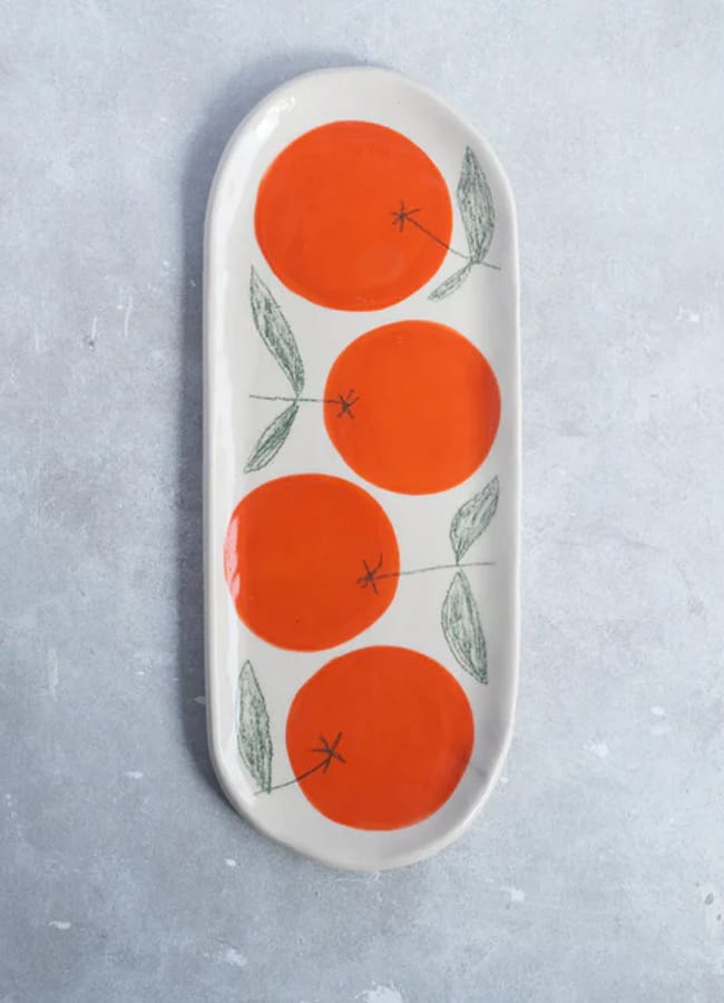 An oval ceramic plate with four large orange circles and green leaves, resembling stylised oranges, sits on a light grey textured surface—a vibrant addition to any foodie homewares collection.