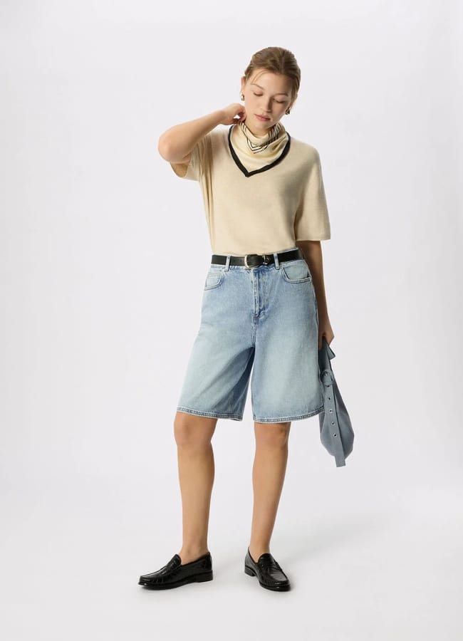 A person stands against a plain background wearing a beige short-sleeve jumper with a black neckline, best denim shorts in light blue, black belt, black loafers, layered necklaces, and holding a blue shirt in one hand.