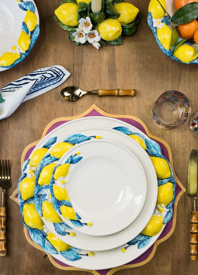 A table set for one with plates decorated with bright yellow lemons and blue leaves, perfect for salad season, features a fork and knife with bamboo-style handles, a glass, serviette, and lemon-themed candle centrepiece on a wooden surface.