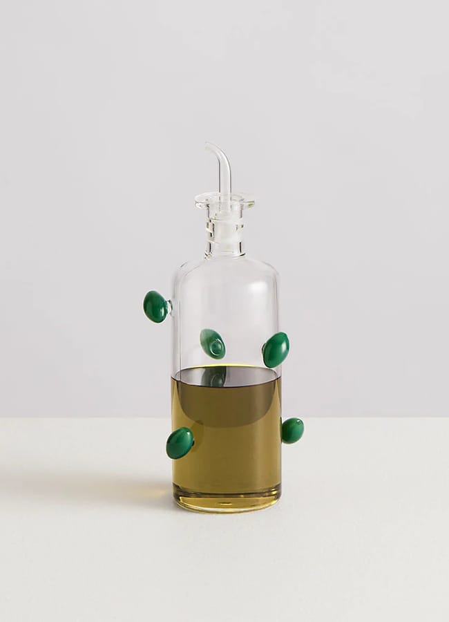 A glass bottle filled with olive oil sits on a white surface, perfect for salad season. Several green olives appear to be floating or attached to the outside of the bottle. The background is plain and light grey.