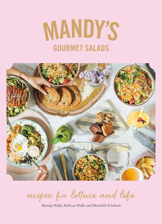A cover of the book Mandy's Gourmet Salads featuring vibrant bowls perfect for salad season, with bread, brownies, avocado, and drinks on a table as hands serve food. The pink background displays enticing text about fresh recipes.