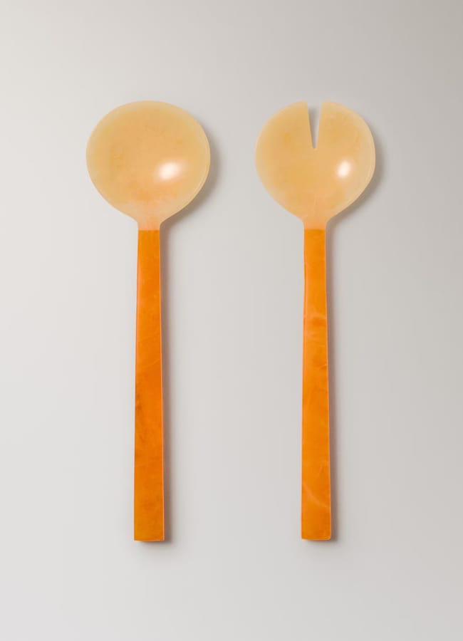 Two salad serving utensils with orange handles and light beige heads, perfect for salad season—one is spoon-shaped and the other fork-shaped with a single slot, placed side by side on a neutral background.
