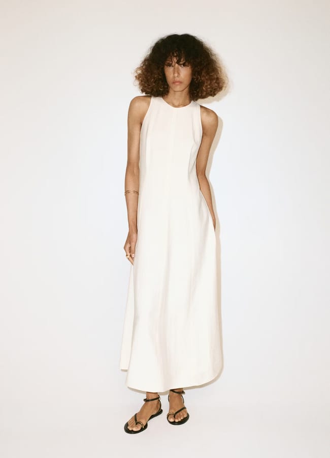 Person with curly hair wears a sleeveless, long, white dress and black sandals, standing against a plain white background. Their arms are relaxed at their sides, embodying effortless high street fashion.