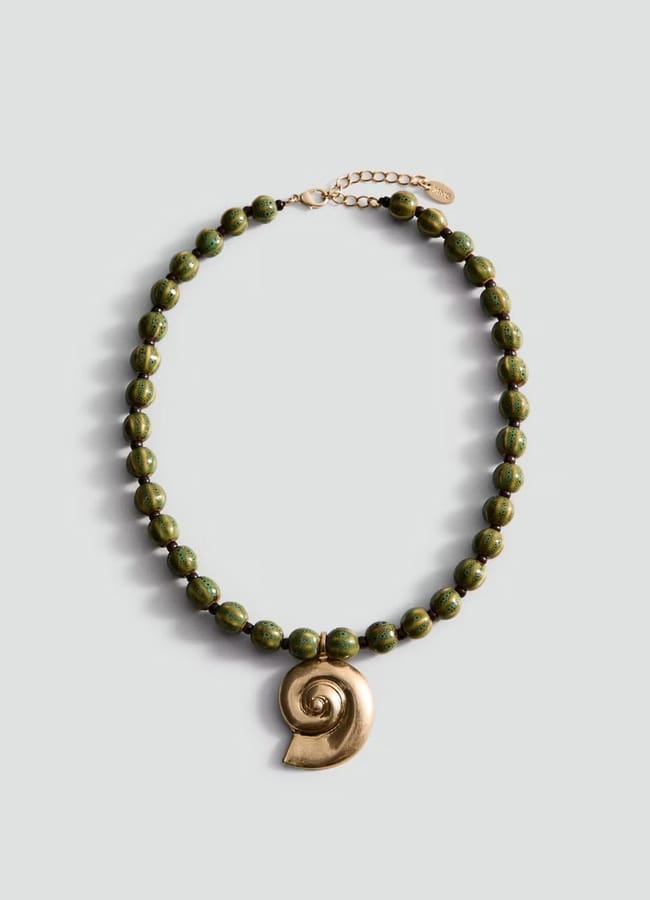 A green beaded necklace with a gold spiral shell pendant, displayed on a light grey background—this unique accessory is perfect inspiration for what to wear in Ibiza.