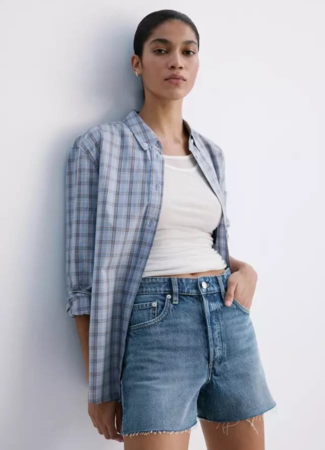 A person stands against a white wall wearing a blue checked button-up shirt over a white vest, paired with the best denim shorts. Their hair is tied back, and they have a relaxed, confident expression.