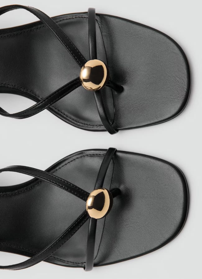 A close-up top view of black strappy sandals with square toes and gold round ornaments on each sandal, set against a light background—perfect for elevating any high street fashion look.