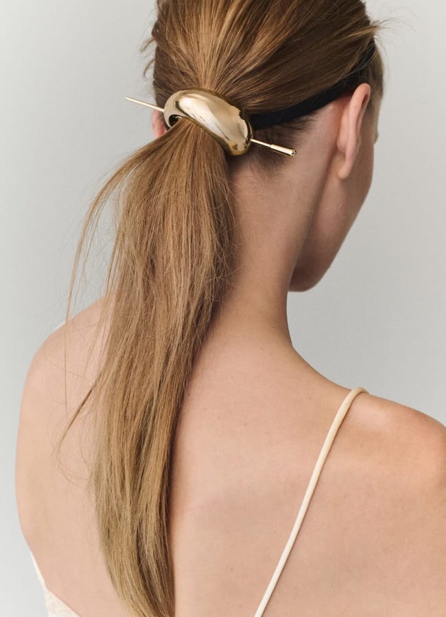 A woman with light brown hair styled in a low ponytail, secured with a shiny gold hair accessory—a chic touch that reflects high street fashion. She is wearing a thin-strapped top and is facing away from the camera.