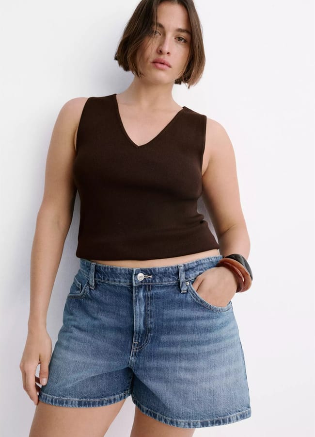A person with short brown hair wears a sleeveless dark brown V-neck top and the best denim shorts, standing against a plain white background with one hand in their pocket.