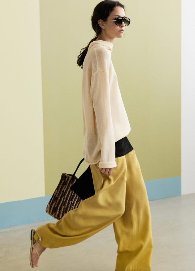 A person embodying high street fashion walks indoors, wearing sunglasses, a sheer beige polo neck, loose mustard yellow trousers, and sandals whilst carrying a woven bag.