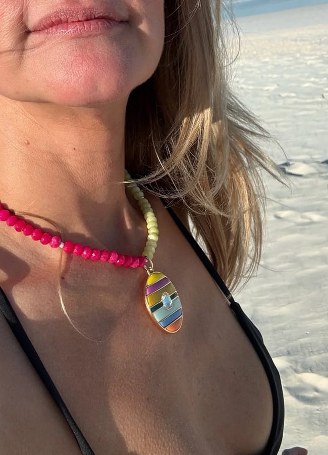 A woman wearing a black bikini top and colourful beach jewellery, including a vibrant beaded necklace with a striped pendant, stands on a sandy beach under bright sunlight.