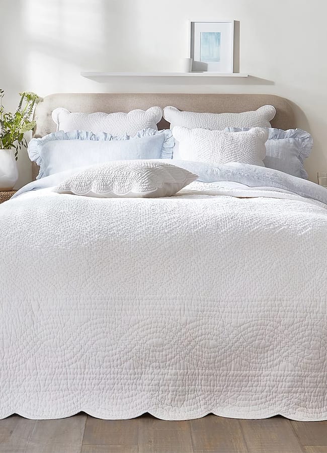 A neatly made bed with white and light blue textured bedding, ruffled pillows, and a scalloped quilt evokes the cosy elegance often found in maximalist hotels. A small vase with green flowers and a framed picture sit on a shelf above the bed.
