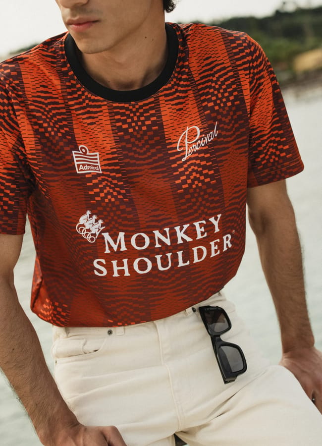 A person wearing a red and black patterned shirt with white logos and text reading “Percival,” “Monkey Shoulder,” and “Admiral” channels football core style, paired with beige trousers and sunglasses clipped to the waistband.